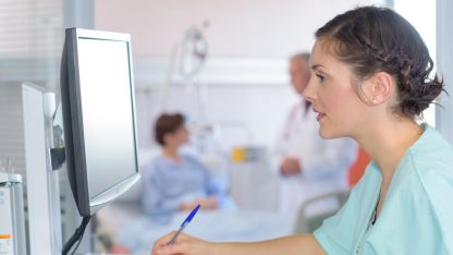 Eine Pflegerin sitzt am Computer und macht Notizen auf einem Block