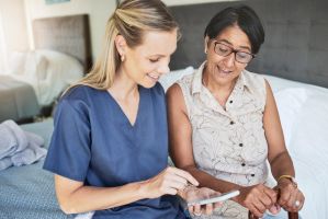 Pflegerin mit sitzt neben einer Patientin am Bett mit Smartphone in der Hand