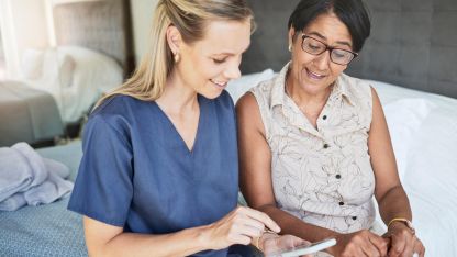 Pflegerin mit sitzt neben einer Patientin am Bett mit Smartphone in der Hand