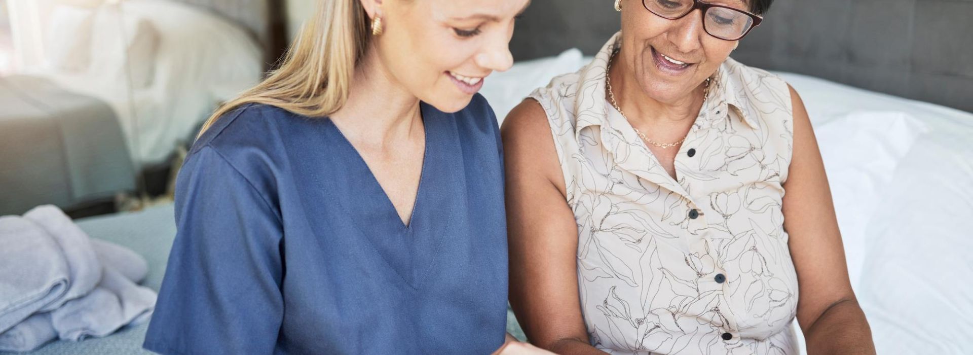 Pflegerin mit sitzt neben einer Patientin am Bett mit Smartphone in der Hand